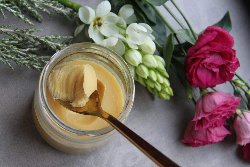 Natural beef tallow in a glass jar on a wooden surface, with a small spoon  – rich in fatty acids and vitamins A, D, E, and K, tallow is gaining popularity in skincare for its skin-replenishing and barrier-supporting properties.